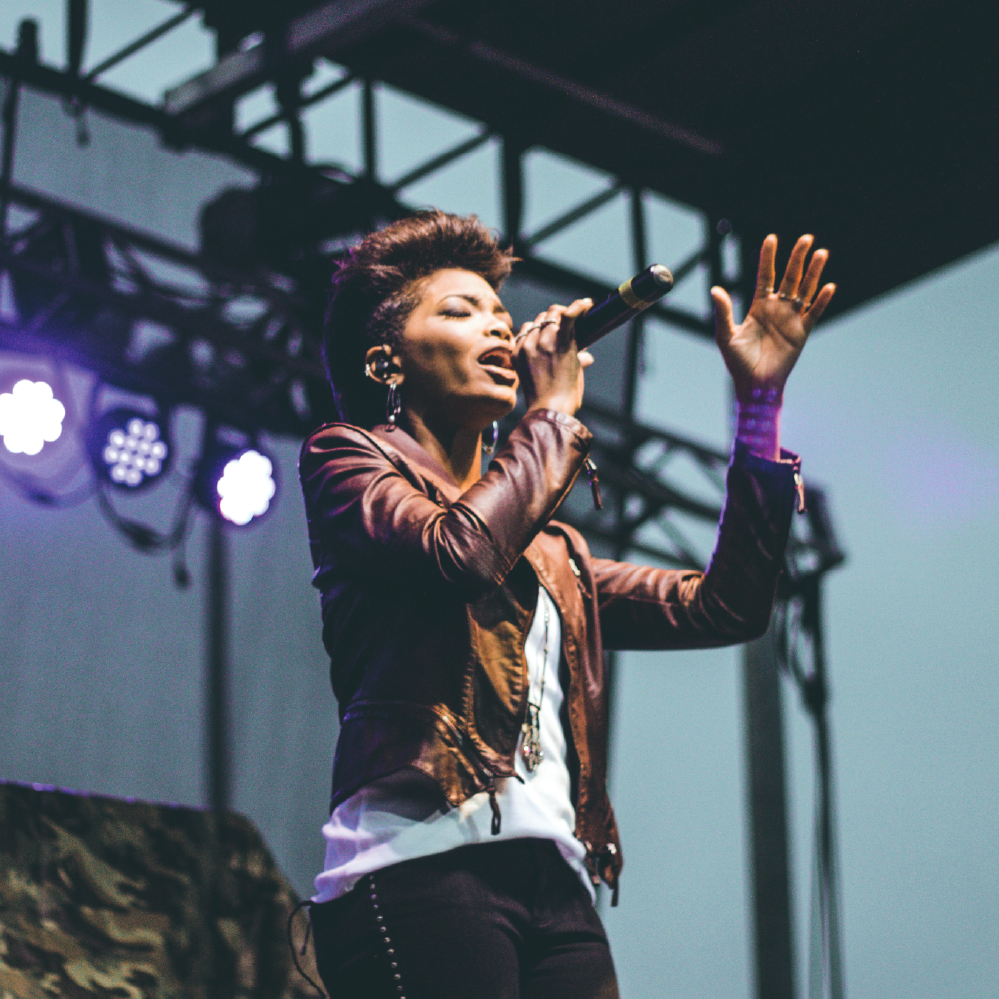 Promotional image for the 2026 New Mexico All-State Vocal Jazz Ensemble with a photo of a woman singing on stage at a jazz festival, with text overlay detailing the ensemble's name and the fact that it is a vocal jazz ensemble.