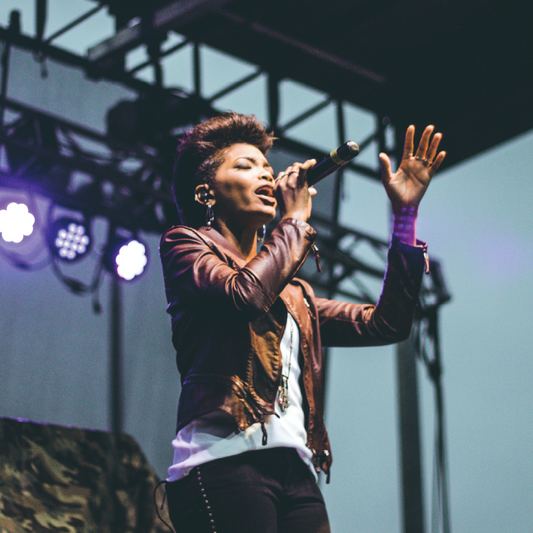Promotional image for the 2026 New Mexico All-State Vocal Jazz Ensemble with a photo of a woman singing on stage at a jazz festival, with text overlay detailing the ensemble's name and the fact that it is a vocal jazz ensemble.